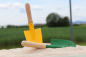 Preview: Zwei Sandschaufeln für Kinder stecken  im Sand eines Spielplatzes. Sie haben einen gelben und grünen Besatz. Dahinter befindet sich eine Wiese.